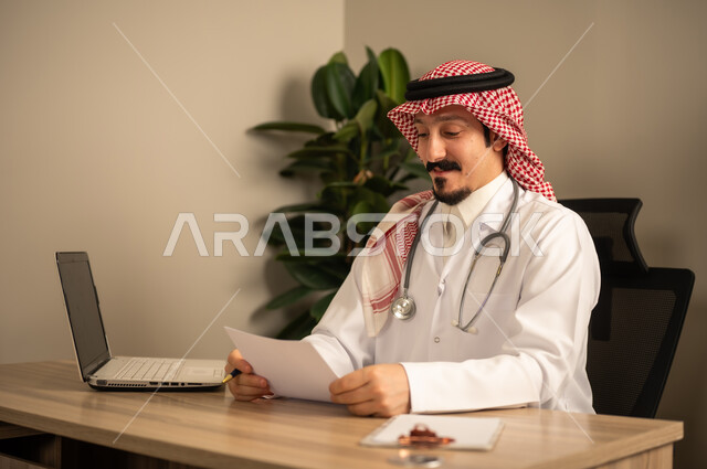قراءة التحاليل والتقارير الطبية، طبيب عربي خليجي سعودي يرتدي المعطف الأبيض يضع السماعات الطبيه حول رقبته يستخدم الحاسوب المحمول، يحمل بيده أوراق وملفات المرضى، العمل في القطاع الصحي في المملكة العربية السعودية، تقديم الرعاية الصحية للمرضى
