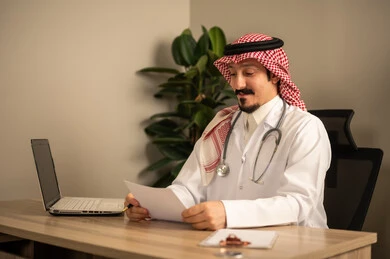 Reading medical analyses and reports, an Arab Gulf Saudi doctor wearing a white coat with a stethoscope around his neck using a laptop, holding patient papers and files in his hand, working in the health sector in the Kingdom of Saudi Arabia, providing healthcare to patients.