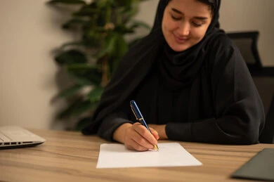 A Saudi Arabian Gulf woman wearing a black abaya and hijab sits in the office, blogging and writing work reports, engaging in office professions and tasks, providing a comfortable and suitable work environment, using a laptop to complete tasks professionally, and employment in Saudi offices and companies.