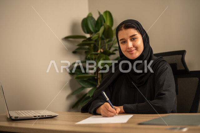 توفير بيئة عمل مريحة ومناسبة، مهن وأعمال مكتبية،امراه عربية خليجية سعودية ترتدي العباءة السوداء والحجاب تجلس في المكتب تقوم بالتدوين وكتابة تقارير العمل ،  استعمال جهاز الحاسوب المحمول لإنجاز المهام باحترافية، التوظيف في المكاتب والشركات السعودية
