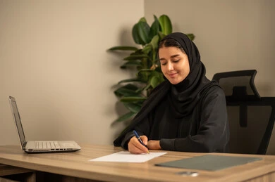 مهن وأعمال مكتبية،  توفير بيئة عمل مريحة ومناسبة، امراه عربية خليجية سعودية ترتدي العباءة السوداء والحجاب تجلس في المكتب تقوم بالتدوين وكتابة تقارير العمل ،  استعمال جهاز الحاسوب المحمول لإنجاز المهام باحترافية، التوظيف في المكاتب والشركات السعودية