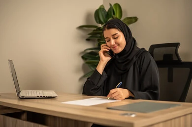 تدوين وكتابة التقارير، مهن وأعمال مكتبية، استخدام الهاتف المحمول، توفير بيئة عمل مريحة ومناسبة، استعمال جهاز الحاسوب المحمول لإنجاز المهام باحترافية، امراه عربية خليجية سعودية ترتدي العباءة السوداء والحجاب تجلس في المكتب تقوم بالعمل عبر اللابتوب، التوظيف في المكاتب والشركات السعودية
