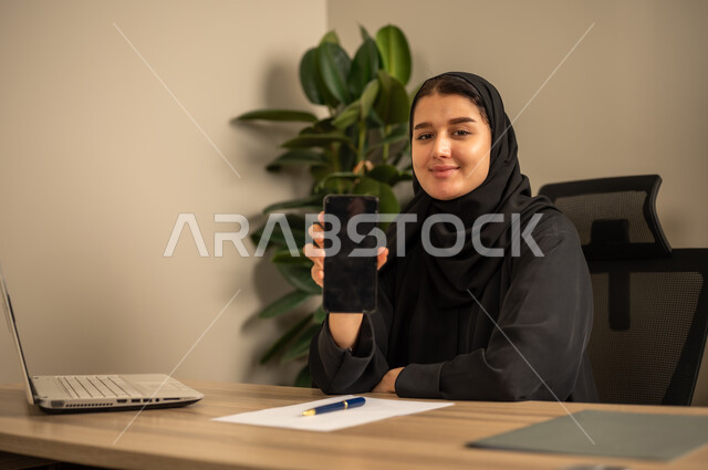 عرض شاشة سوداء فارغة، مهن وأعمال مكتبية، استخدام الهاتف المحمول، توفير بيئة عمل مريحة ومناسبة، استعمال جهاز الحاسوب المحمول لإنجاز المهام باحترافية، امراه عربية خليجية سعودية ترتدي العباءة السوداء والحجاب  تجلس في المكتب تقوم بالعمل عبر اللابتوب، التوظيف في المكاتب والشركات السعودية