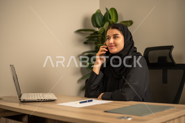 استخدام الهاتف المحمول، تدوين وكتابة التقارير، مهن وأعمال مكتبية،توفير بيئة عمل مريحة ومناسبة، استعمال جهاز الحاسوب المحمول لإنجاز المهام باحترافية، امراه عربية خليجية سعودية ترتدي العباءة السوداء والحجاب تجلس في المكتب تقوم بالعمل عبر اللابتوب، التوظيف في المكاتب والشركات السعودية