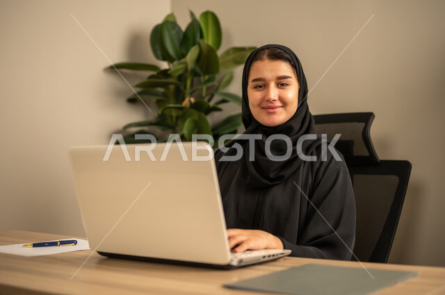 استخدام جهاز الحاسوب المحمول لإنجاز المهام باحترافية،التوظيف في المكاتب والشركات السعودية، توفير بيئة عمل مريحة ومناسبة،  سيدة عربية خليجية سعودية يرتدي العباءة السوداء والحجاب تقوم بالعمل عبر اللابتوب داخل المكتب