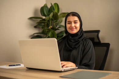 Using a laptop to complete tasks professionally, employment in Saudi offices and companies, providing a comfortable and suitable work environment, an Arab Gulf Saudi woman wearing a black abaya and hijab is working on a laptop inside the office.
