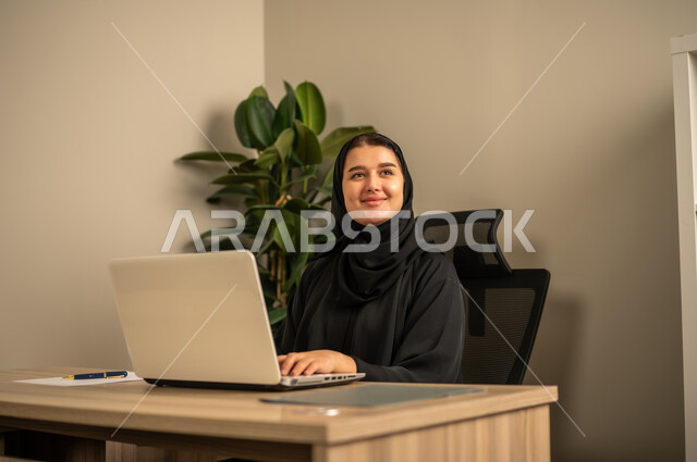 Providing a comfortable and suitable work environment, employment in Saudi offices and companies, using a laptop to complete tasks professionally, an Arab Gulf Saudi woman wearing a black abaya and hijab working on a laptop inside the office.