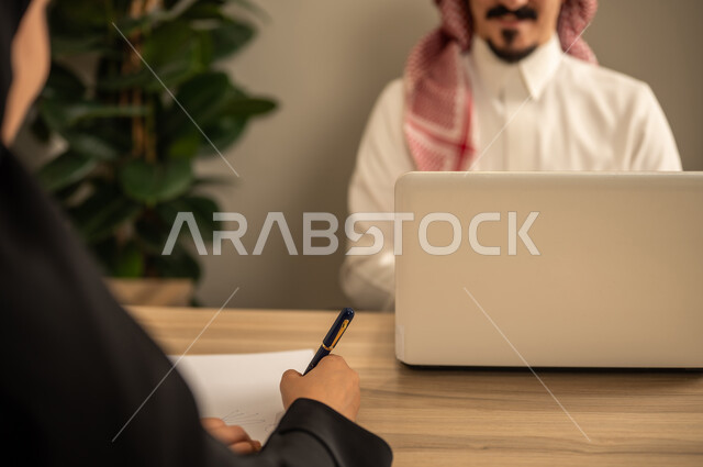 Task management and division, using a laptop, two Arab Gulf Saudi employees conducting a business meeting inside the conference room at the company headquarters, integrating modern technology with office work, studying work papers, developing projects and investments.