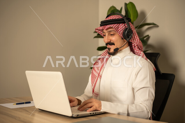 Answering customer questions, using modern technologies, a Gulf Arab Saudi man wearing traditional dress and a ghutrah working in customer service wearing a headset and using a laptop, the concept of inquiries and questions, monitoring and developing work.