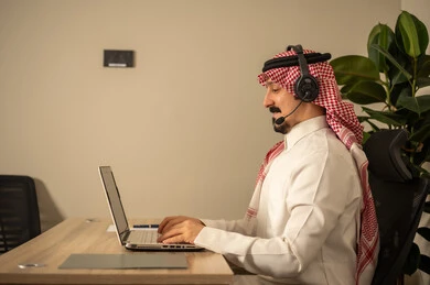 مفهوم الاستعلامات والاستفسارات، الاجابه على اسئلة العملاء، استخدام التقنيات الحديثة، رجل عربي خليجي سعودي يرتدى الثوب التقليدي والشماغ يعمل في خدمة العملاء ترتدي سماعة الاتصال يستخدم الحاسوب المحمول،متابعة وتطوير العمل