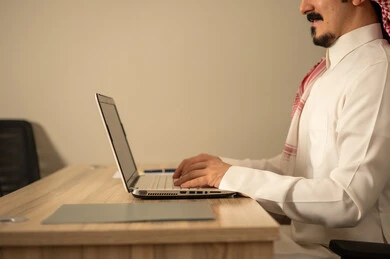 Employment in Saudi offices and companies, providing a comfortable and suitable work environment, using a laptop to complete tasks professionally, an Arab Gulf Saudi man wearing traditional thobe and ghutrah working on a laptop inside the office.
