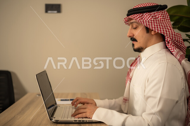 دمج العمل مع التكنولوجيا المتطورة، التوظيف في المكاتب والشركات السعودية، توفير بيئة عمل مريحة ومناسبة،  استخدام جهاز الحاسوب المحمول لإنجاز المهام باحترافية، رجل عربي خليجي سعودي يرتدي الثوب والشماغ التقليدي يقوم بالعمل عبر اللابتوب داخل المكتب
