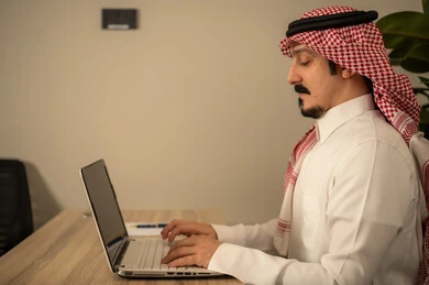 Integrating work with advanced technology, employment in Saudi offices and companies, providing a comfortable and suitable work environment, using a laptop to accomplish tasks professionally, an Arab Gulf Saudi man wearing traditional thobe and ghutrah working on a laptop inside the office.