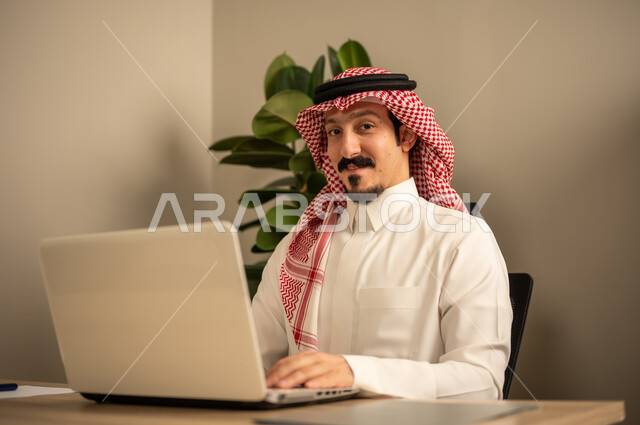 استخدام جهاز الحاسوب المحمول لإنجاز المهام باحترافية،التوظيف في المكاتب والشركات السعودية، توفير بيئة عمل مريحة ومناسبة،   رجل عربي خليجي سعودي يرتدي الثوب والشماغ التقليدي يقوم بالعمل عبر اللابتوب داخل المكتب