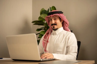 استخدام جهاز الحاسوب المحمول لإنجاز المهام باحترافية،التوظيف في المكاتب والشركات السعودية، توفير بيئة عمل مريحة ومناسبة،   رجل عربي خليجي سعودي يرتدي الثوب والشماغ التقليدي يقوم بالعمل عبر اللابتوب داخل المكتب