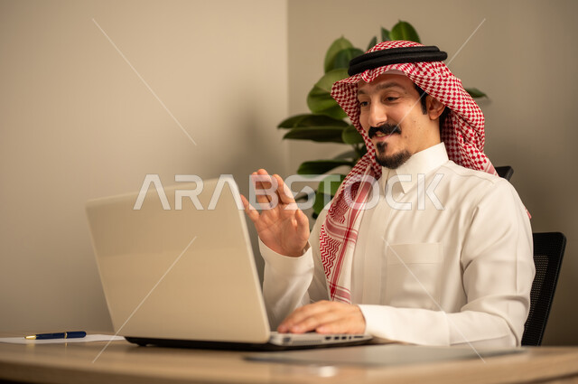 Providing a comfortable and suitable work environment, employment in Saudi offices and companies, using a laptop to complete tasks professionally, an Arab Gulf Saudi man wearing traditional thobe and ghutrah working on a laptop inside the office.