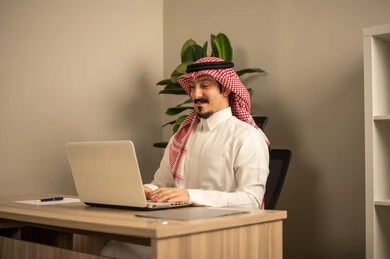 التوظيف في المكاتب والشركات السعودية، توفير بيئة عمل مريحة ومناسبة،  استخدام جهاز الحاسوب المحمول لإنجاز المهام باحترافية، رجل عربي خليجي سعودي يرتدي الثوب والشماغ التقليدي يقوم بالعمل عبر اللابتوب داخل المكتب