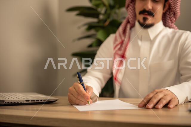 تدوين وكتابة التقارير، مهن وأعمال مكتبية، استخدام الهاتف المحمول، توفير بيئة عمل مريحة ومناسبة، استعمال جهاز الحاسوب المحمول لإنجاز المهام باحترافية، رجل عربي خليجي سعودي يرتدي الثوب والشماغ التقليدي يجلس في المكتب يقوم بالعمل عبر اللابتوب، التوظيف في المكاتب والشركات السعودية