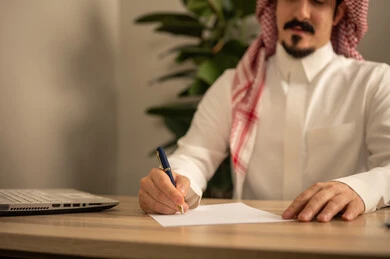 Writing and documenting reports, office jobs and tasks, using mobile phones, providing a comfortable and suitable work environment, using a laptop to complete tasks professionally, an Arab Gulf Saudi man wearing traditional thobe and ghutrah sitting in the office working on the laptop, employment in Saudi offices and companies.