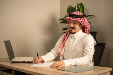 تدوين وكتابة التقارير، مهن وأعمال مكتبية، استخدام الهاتف المحمول، توفير بيئة عمل مريحة ومناسبة، استعمال جهاز الحاسوب المحمول لإنجاز المهام باحترافية، رجل عربي خليجي سعودي يرتدي الثوب والشماغ التقليدي يجلس في المكتب يقوم بالعمل عبر اللابتوب، التوظيف في المكاتب والشركات السعودية