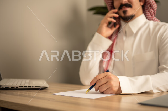 Making a phone call, taking notes and writing reports, office jobs and tasks, using a mobile phone, providing a comfortable and suitable work environment, using a laptop to complete tasks professionally, an Arab Gulf Saudi man wearing traditional thobe and ghutrah sitting in the office working on the laptop, employment in Saudi offices and companies.