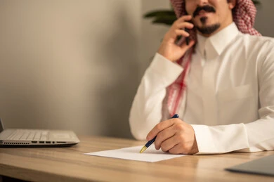 Making a phone call, taking notes and writing reports, office jobs and tasks, using a mobile phone, providing a comfortable and suitable work environment, using a laptop to complete tasks professionally, an Arab Gulf Saudi man wearing traditional thobe and ghutrah sitting in the office working on the laptop, employment in Saudi offices and companies.