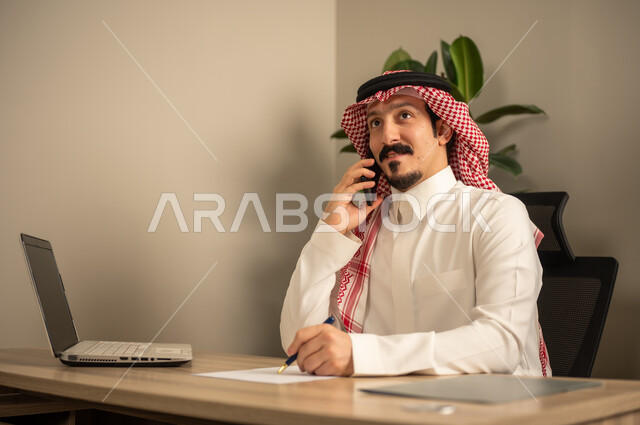 توفير بيئة عمل مريحة ومناسبة،تدوين وكتابة التقارير، مهن وأعمال مكتبية، استخدام الهاتف المحمول، استعمال جهاز الحاسوب المحمول لإنجاز المهام باحترافية، رجل عربي خليجي سعودي يرتدي الثوب والشماغ التقليدي يجلس في المكتب يقوم بالعمل عبر اللابتوب، التوظيف في المكاتب والشركات السعودية