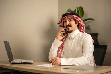 توفير بيئة عمل مريحة ومناسبة،تدوين وكتابة التقارير، مهن وأعمال مكتبية، استخدام الهاتف المحمول، استعمال جهاز الحاسوب المحمول لإنجاز المهام باحترافية، رجل عربي خليجي سعودي يرتدي الثوب والشماغ التقليدي يجلس في المكتب يقوم بالعمل عبر اللابتوب، التوظيف في المكاتب والشركات السعودية