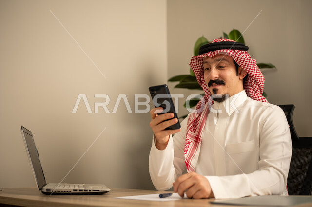 استخدام الهاتف المحمول، تدوين وكتابة التقارير، مهن وأعمال مكتبية، توفير بيئة عمل مريحة ومناسبة، استعمال جهاز الحاسوب المحمول لإنجاز المهام باحترافية، رجل عربي خليجي سعودي يرتدي الثوب والشماغ التقليدي يجلس في المكتب يقوم بالعمل عبر اللابتوب، التوظيف في المكاتب والشركات السعودية