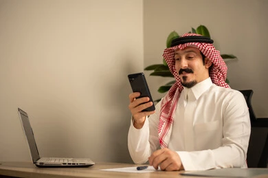 استخدام الهاتف المحمول، تدوين وكتابة التقارير، مهن وأعمال مكتبية، توفير بيئة عمل مريحة ومناسبة، استعمال جهاز الحاسوب المحمول لإنجاز المهام باحترافية، رجل عربي خليجي سعودي يرتدي الثوب والشماغ التقليدي يجلس في المكتب يقوم بالعمل عبر اللابتوب، التوظيف في المكاتب والشركات السعودية