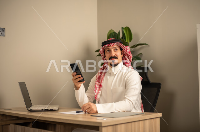 مهن وأعمال مكتبية، تدوين وكتابة التقارير، استخدام الهاتف المحمول، توفير بيئة عمل مريحة ومناسبة، استعمال جهاز الحاسوب المحمول لإنجاز المهام باحترافية، رجل عربي خليجي سعودي يرتدي الثوب والشماغ التقليدي يجلس في المكتب يقوم بالعمل عبر اللابتوب، التوظيف في المكاتب والشركات السعودية