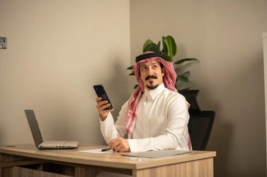 مهن وأعمال مكتبية، تدوين وكتابة التقارير، استخدام الهاتف المحمول، توفير بيئة عمل مريحة ومناسبة، استعمال جهاز الحاسوب المحمول لإنجاز المهام باحترافية، رجل عربي خليجي سعودي يرتدي الثوب والشماغ التقليدي يجلس في المكتب يقوم بالعمل عبر اللابتوب، التوظيف في المكاتب والشركات السعودية