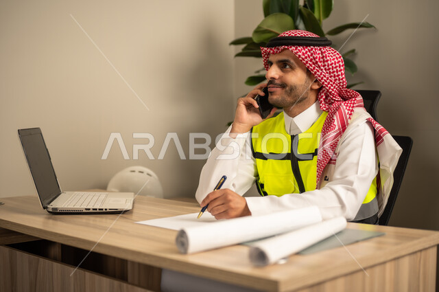 Project auditing and review, engineering tools for completing the construction plan drawing, integrating work with technology and tech, a Saudi Gulf Arab engineer wearing a traditional headscarf and thobe working on a laptop while holding a mobile phone and completing the plan.