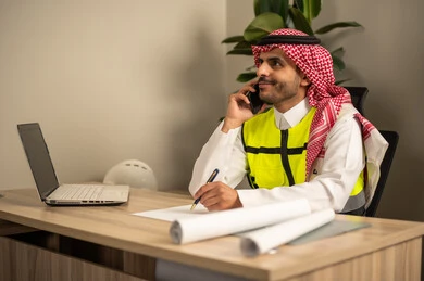 Project auditing and review, engineering tools for completing the construction plan drawing, integrating work with technology and tech, a Saudi Gulf Arab engineer wearing a traditional headscarf and thobe working on a laptop while holding a mobile phone and completing the plan. Project auditing and review, engineering tools for completing the construction plan drawing, integrating work with technology and tech, a Saudi Gulf Arab engineer wearing a traditional headscarf and thobe working on a laptop while holding a mobile phone and completing the plan.