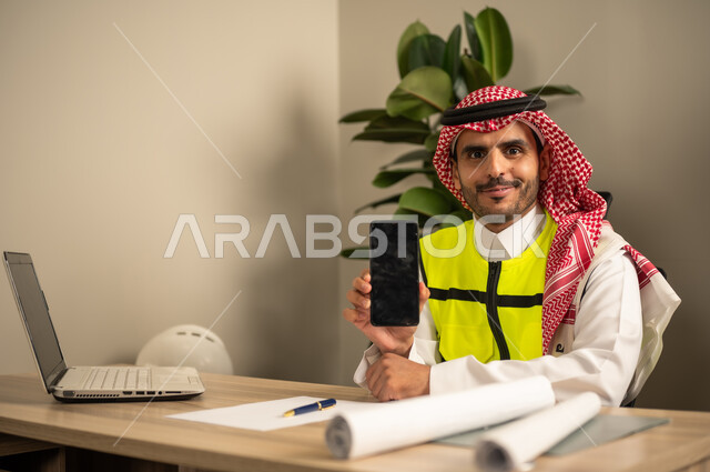 Project auditing and review, engineering tools for completing the construction plan drawing, integrating work with technology and tech, a Saudi Gulf Arab engineer wearing a traditional headscarf and thobe working on a laptop while holding a mobile phone and completing the plan.