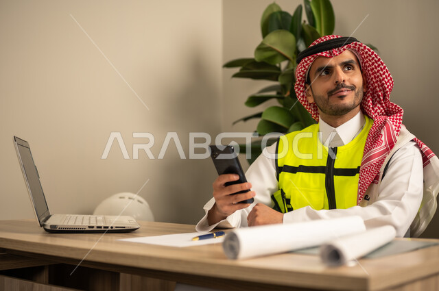 Engineering tools to complete the construction plan drawing, project auditing and reviewing, integrating work with technology and tech, a Saudi Gulf Arab engineer wearing a ghutrah and traditional thobe working on a laptop while holding a mobile phone and completing the plan.
