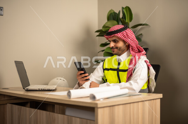تدقيق المشاريع ومراجعتها، أدوات هندسية لإكمال رسم مخطط البناء، دمج العمل بالتقنية والتكنولوجيا, مهندس عربي خليجي سعودي يرتدي الشماغ والثوب التقليدي يعمل على جهاز اللابتوب المحمول يمسك بيده الهاتف المحمول ويستكمل المخطط