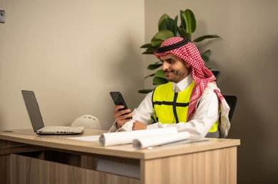 Project auditing and review, engineering tools for completing the construction plan drawing, integrating work with technology and tech, a Saudi Gulf Arab engineer wearing a traditional headscarf and thobe working on a laptop while holding a mobile phone and completing the plan.