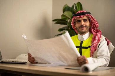 Architectural designs and plan auditing, development and evolution of work in the engineering field, a Saudi Gulf Arab engineer wearing a safety coat holding a work plan, engineering project management, Saudi professions and jobs.