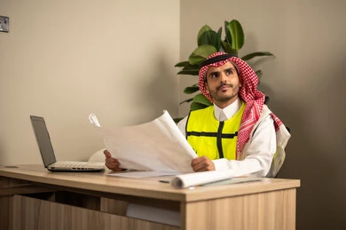 A Saudi Gulf Arab engineer wearing a safety coat is following plans on a laptop while holding a work plan in his hand, reviewing and reading blueprints, working in the field of structural engineering, using technical devices in engineering professions, and supervising the progress of construction work. A Saudi Gulf Arab engineer wearing a safety coat is following plans on a laptop while holding a work plan in his hand, reviewing and reading blueprints, working in the field of structural engineering, using technical devices in engineering professions, and supervising the progress of construction work.