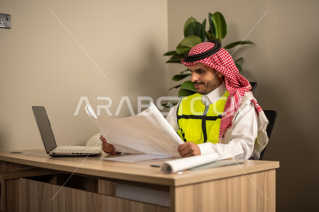 تدقيق وقراءة المخططات، العمل في مجال الهندسة الإنشائية،استخدام الأجهزة التقنية في المهن الهندسية، الإشراف على سير أعمال البناء،مهندس عربي خليجي سعودي يرتدي معطف الحماية يقوم بمتابعة الخطط عبر جهاز اللابتوب يمسك بيده مخطط عمل