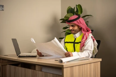 Reviewing and reading plans, working in the field of structural engineering, using technical devices in engineering professions, supervising the progress of construction work, an Arab Gulf Saudi engineer wearing a safety coat is following up on plans via a laptop while holding a work plan in his hand. Reviewing and reading plans, working in the field of structural engineering, using technical devices in engineering professions, supervising the progress of construction work, an Arab Gulf Saudi engineer wearing a safety coat is following up on plans via a laptop while holding a work plan in his hand.