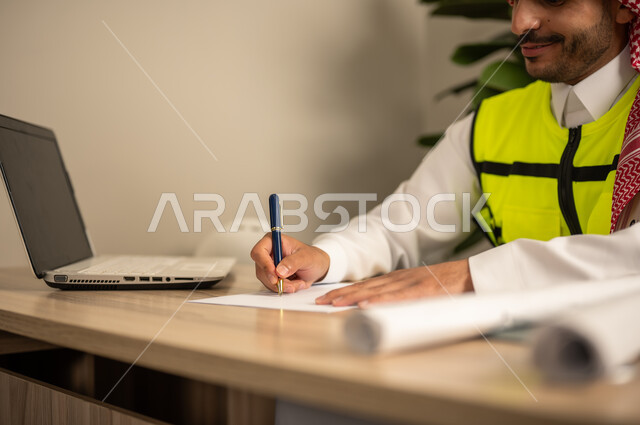Supervising the progress of construction work, documenting architectural project plans, working in the field of structural engineering, using technical devices in engineering professions, an Arab Gulf Saudi engineer wearing a safety coat is following up on plans via a laptop and writing work reports.