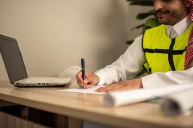 Supervising the progress of construction work, documenting architectural project plans, working in the field of structural engineering, using technical devices in engineering professions, an Arab Gulf Saudi engineer wearing a safety coat is following up on plans via a laptop and writing work reports.