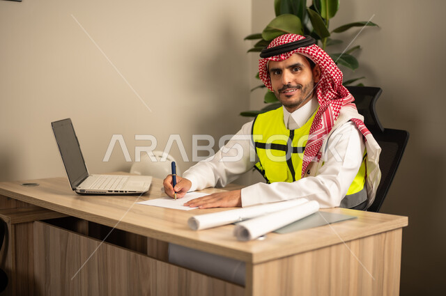 مهندس عربي خليجي سعودي يرتدي معطف الحماية يقوم بمتابعة الخطط عبر جهاز اللابتوب وكتابة تقارير العمل، العمل في مجال الهندسة الإنشائية ، تدوين خطط المشاريع المعمارية، استخدام الأجهزة التقنية في المهن الهندسية، الإشراف على سير أعمال البناء