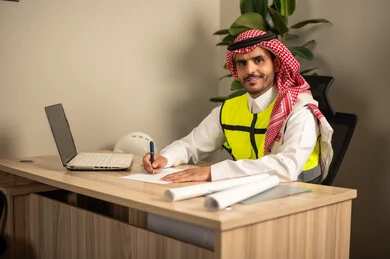 A Saudi Gulf Arab engineer wearing a safety coat is following up on plans via a laptop and writing work reports, working in the field of structural engineering, documenting architectural project plans, using technical devices in engineering professions, and supervising the progress of construction work.