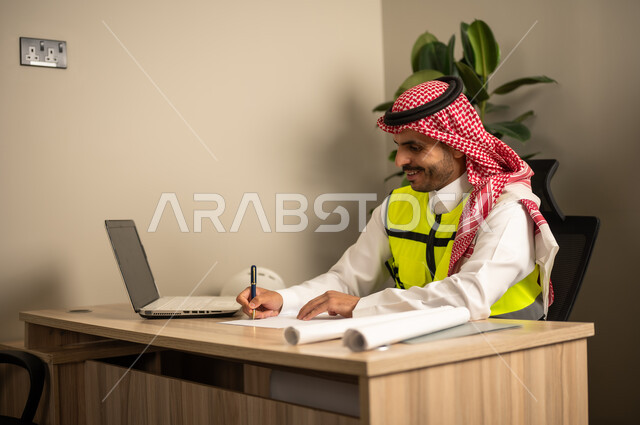 تدوين خطط المشاريع المعمارية، العمل في مجال الهندسة الإنشائية،استخدام الأجهزة التقنية في المهن الهندسية، الإشراف على سير أعمال البناء،مهندس عربي خليجي سعودي يرتدي معطف الحماية يقوم بمتابعة الخطط عبر جهاز اللابتوب وكتابة تقارير العمل