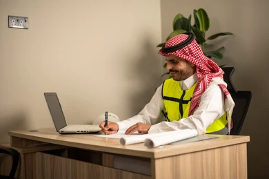 تدوين خطط المشاريع المعمارية، العمل في مجال الهندسة الإنشائية،استخدام الأجهزة التقنية في المهن الهندسية، الإشراف على سير أعمال البناء،مهندس عربي خليجي سعودي يرتدي معطف الحماية يقوم بمتابعة الخطط عبر جهاز اللابتوب وكتابة تقارير العمل