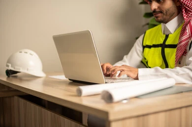 Working in the field of structural engineering, using technical devices in engineering professions, supervising the progress of construction work, following up on the implementation of architectural projects, a Saudi Gulf Arab engineer wearing a safety coat is monitoring plans at the work site via a laptop.