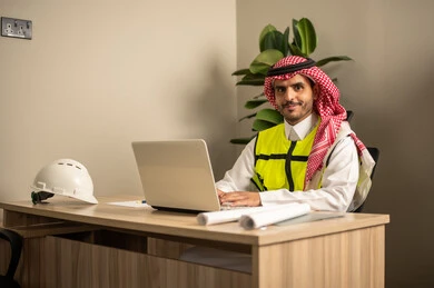 Supervising the progress of construction work, using technical devices in engineering professions, working in the field of structural engineering, monitoring the implementation of architectural projects, an Arab Gulf Saudi engineer wearing a safety coat is following up on plans at the work site via a laptop.